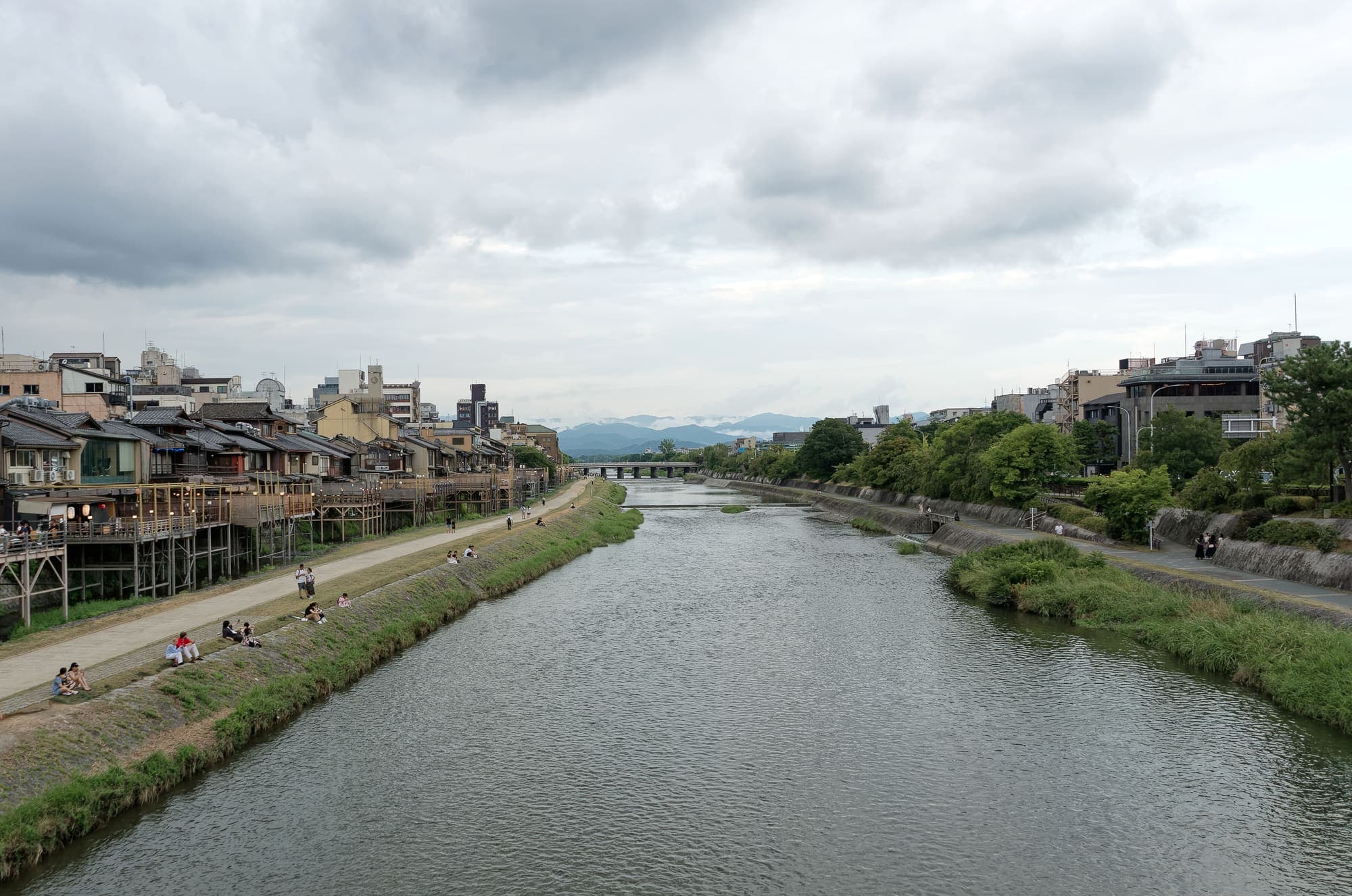 The Kamo River flows north between grassy banks and Pontocho's riverside terraces under an overcast sky, with mountains in the distance.