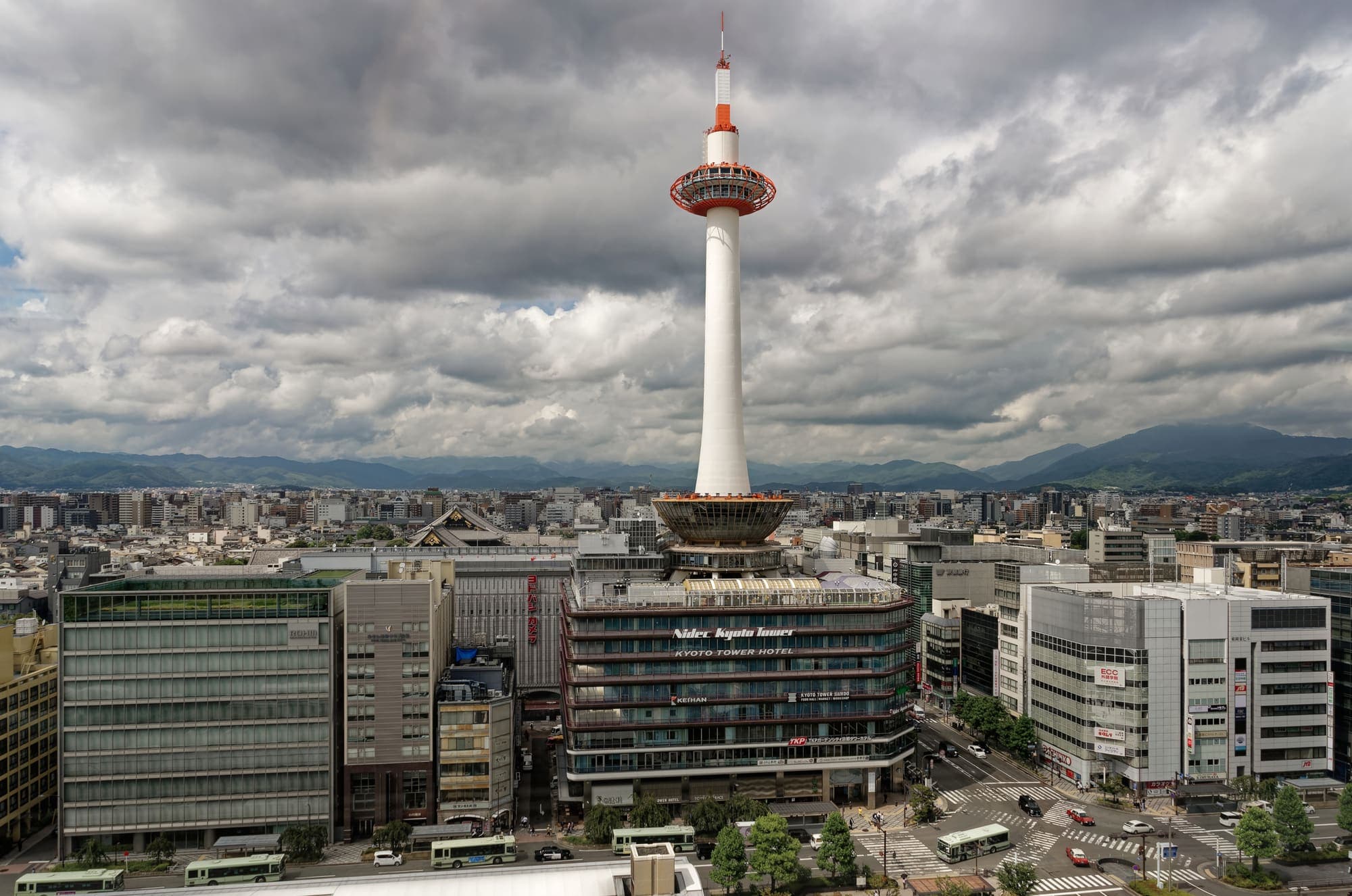 Kyoto Tower rises above office blocks in front of Kyoto Station under a dramatic overcast sky, with mountains on the horizon.